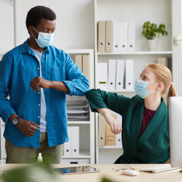 Building employee morale - Seated woman wearing mask bumps elbows to greet man standing next to her, also wearing mask