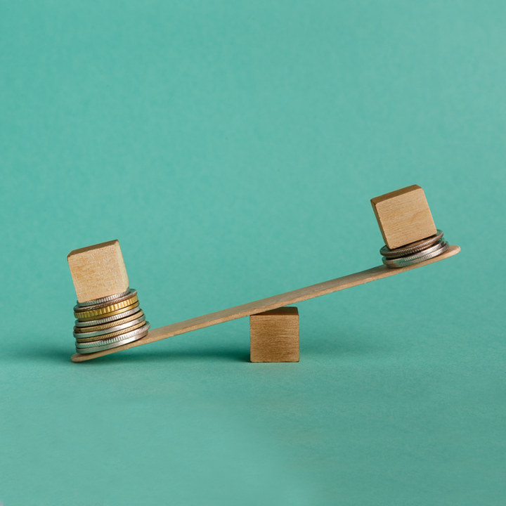 Personal biases - Blue background. Two wooden cubes sitting on piles of coins on opposite ends of a teeter totter. The left cube is the one on the ground because it has more coins underneath. 